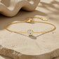 A delicate gold chain bracelet with a single bezel-set round clear gemstone, displayed on a textured beige stone surface with soft shadows and a draped fabric in the background. The tag on the clasp reads 14KT.