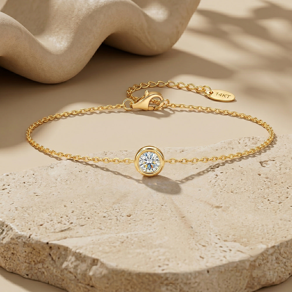 A delicate gold chain bracelet with a single bezel-set round clear gemstone, displayed on a textured beige stone surface with soft shadows and a draped fabric in the background. The tag on the clasp reads 14KT.