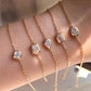 Four delicate gold chain bracelets with clear gemstone settings worn on a person's wrist. The bracelets feature different stone shapes: round, rectangular, teardrop, and marquise, with one bracelet ending in a small cross charm.