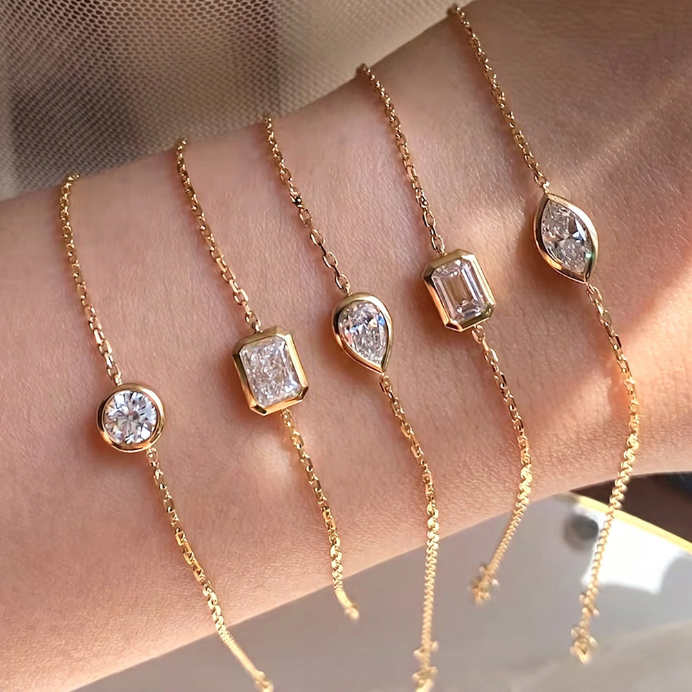 Four delicate gold chain bracelets with clear gemstone settings worn on a person's wrist. The bracelets feature different stone shapes: round, rectangular, teardrop, and marquise, with one bracelet ending in a small cross charm.