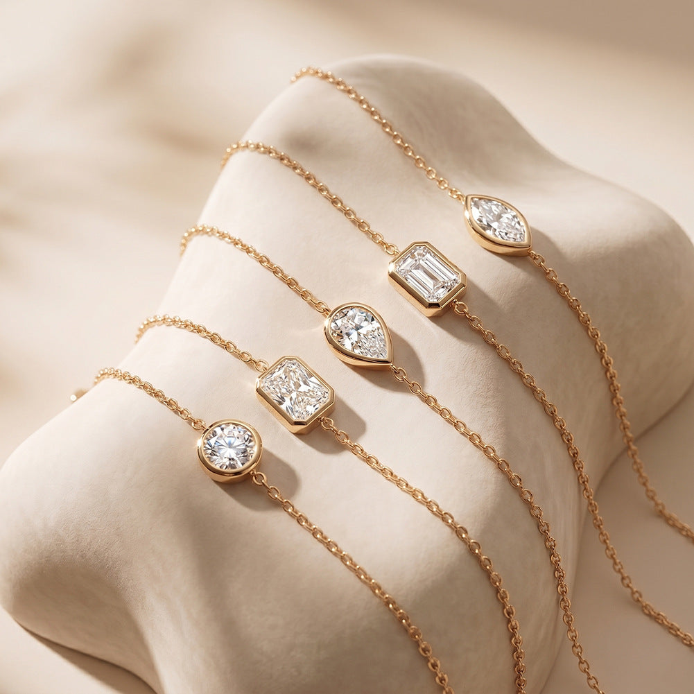 Five delicate gold chain bracelets with different clear gemstone pendants, arranged on a beige fabric display stand.
