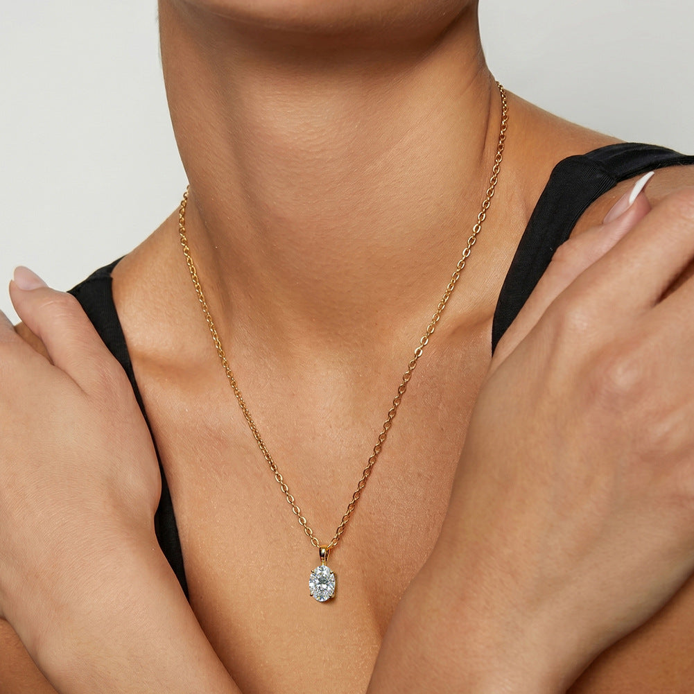 A close-up of a model wearing a delicate gold chain necklace with a small teardrop-shaped clear gemstone pendant, styled with a black top against a neutral light gray background.
