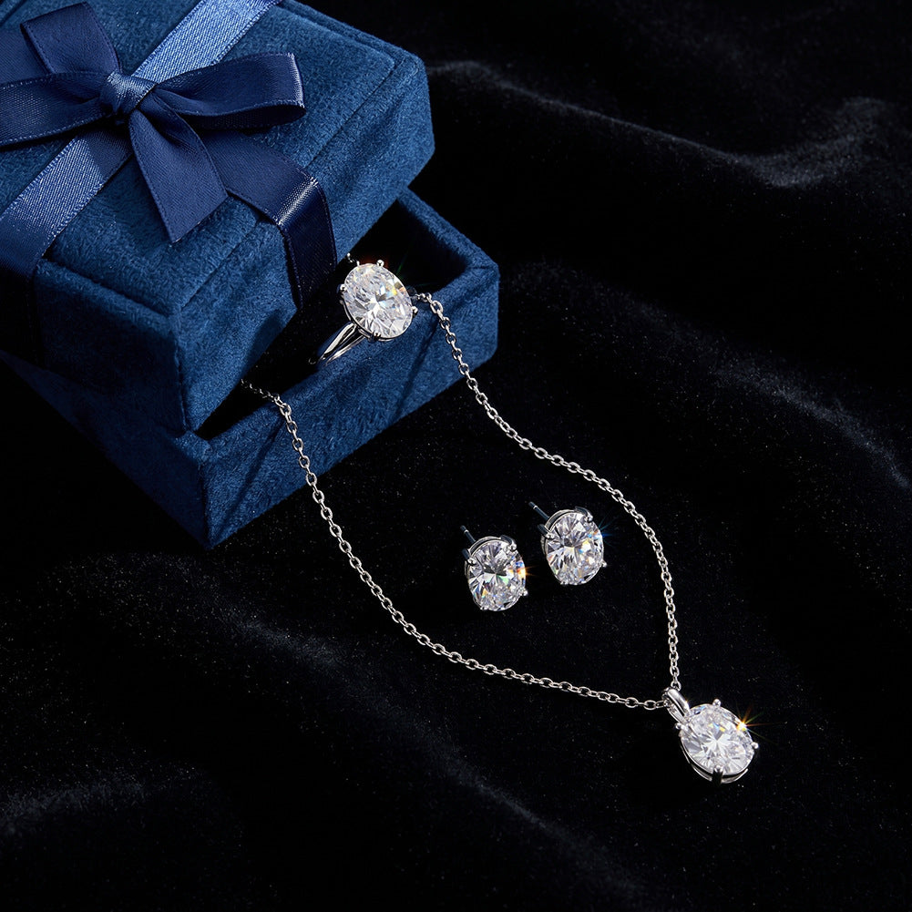A silver-tone jewelry set with a ring, necklace, and earrings, each featuring a clear oval gemstone, displayed on a dark velvet surface next to an open blue velvet gift box with a navy ribbon.