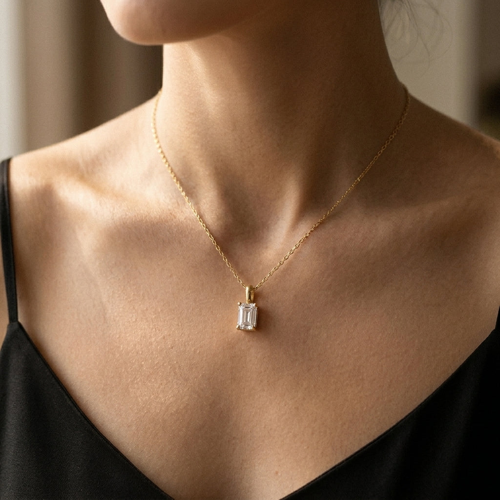 A close-up of a model wearing a delicate gold chain necklace with a rectangular clear baguette-cut gemstone pendant, styled with a black top.