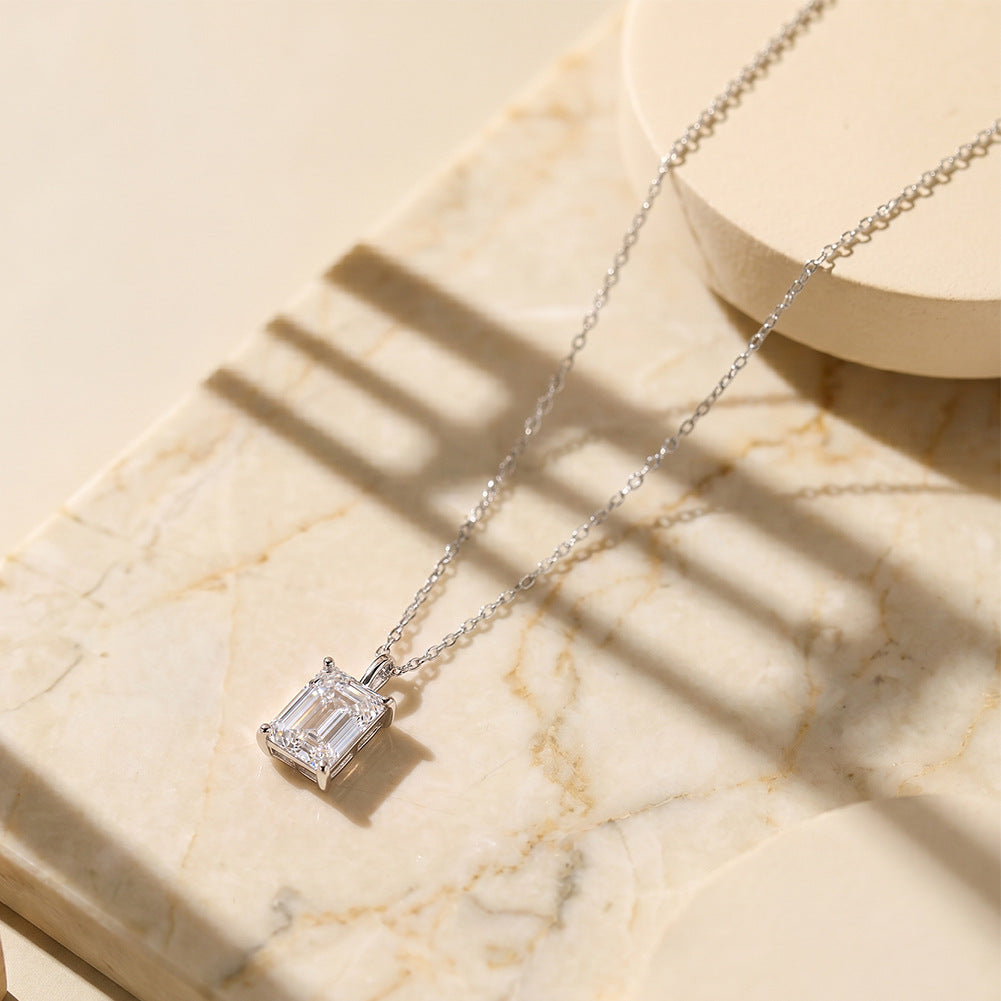 A silver necklace with a rectangular clear gemstone pendant, displayed on a beige marble surface with soft shadows.