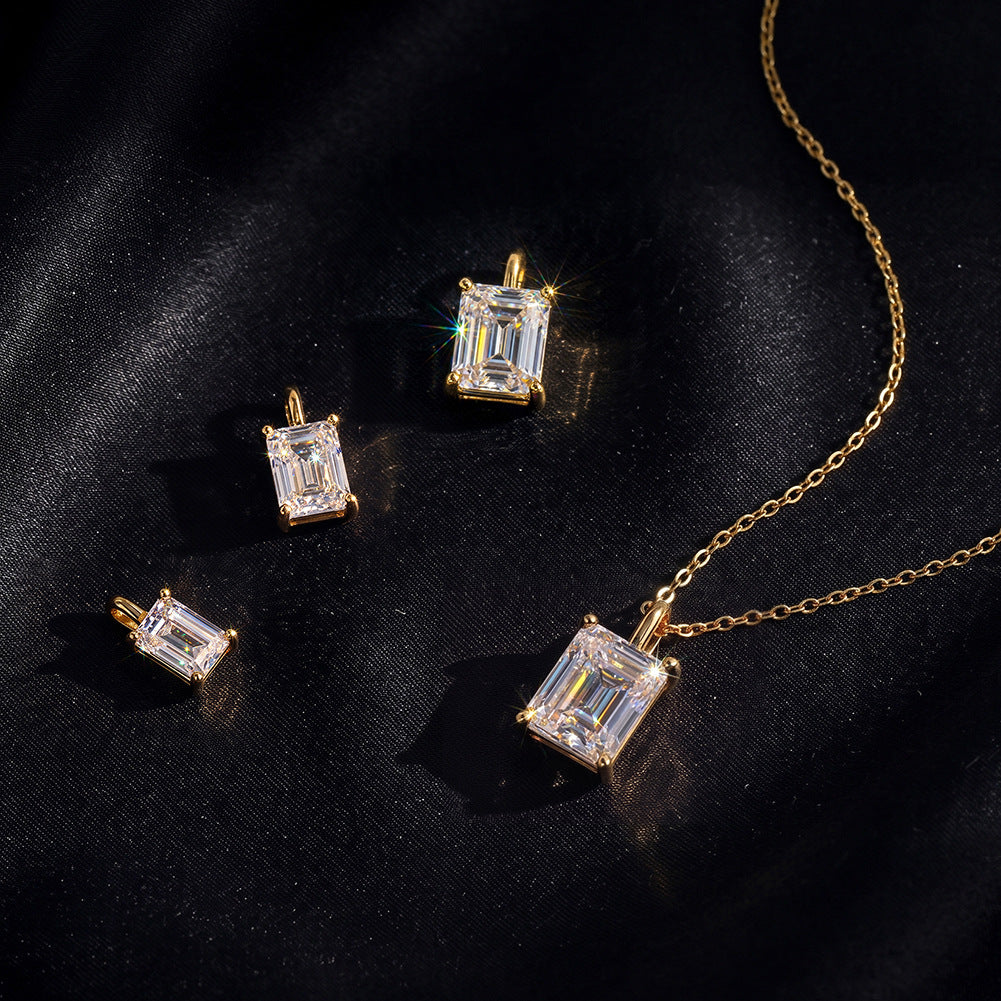 Gold jewelry set featuring an emerald-cut clear gemstone pendant necklace, matching stud earrings, and a smaller rectangular pendant, arranged on a dark black fabric background.
