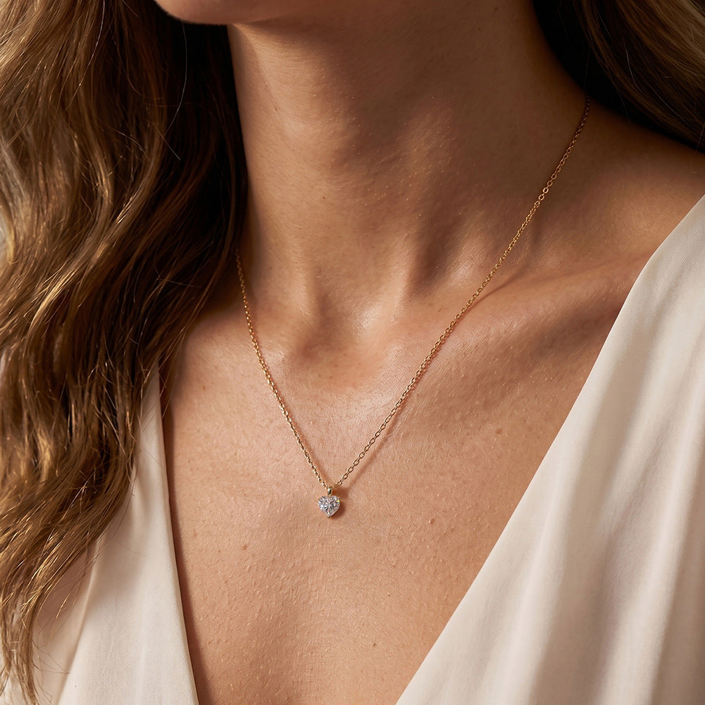 A close-up of a woman's neck and chest wearing a delicate gold chain necklace with a small heart-shaped clear gemstone pendant.