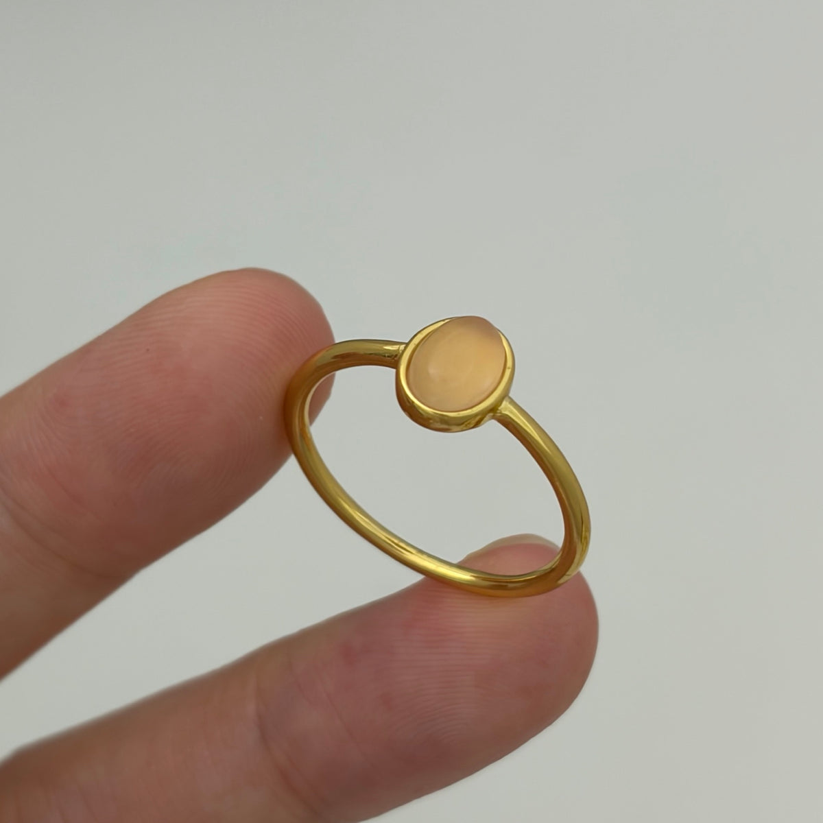 A delicate gold ring with a small, smooth oval cabochon stone held between two fingers against a plain light gray background.
