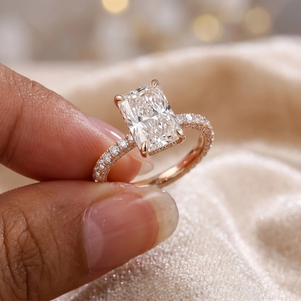 Hand holding 18k yellow gold 2ct radiant cut lab diamond pave engagement ring on champagne satin background