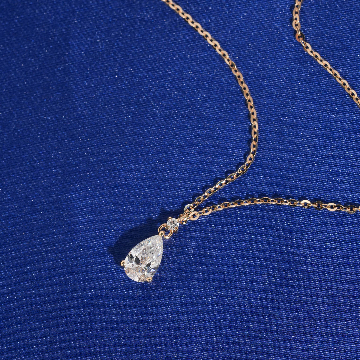 Angle view of 18k rose gold 1 carat pear cut lab-grown diamond clavicle pendant necklace on blue fabric