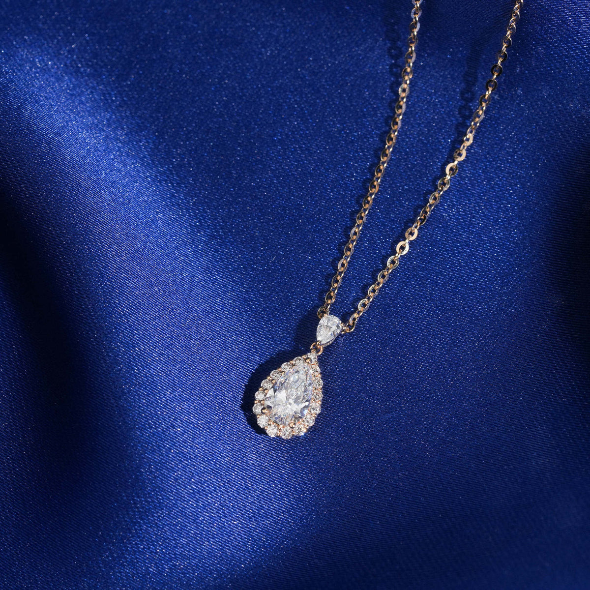 Angle view of 18k rose gold 1 ct pear halo lab-grown diamond clavicle pendant necklace on blue fabric