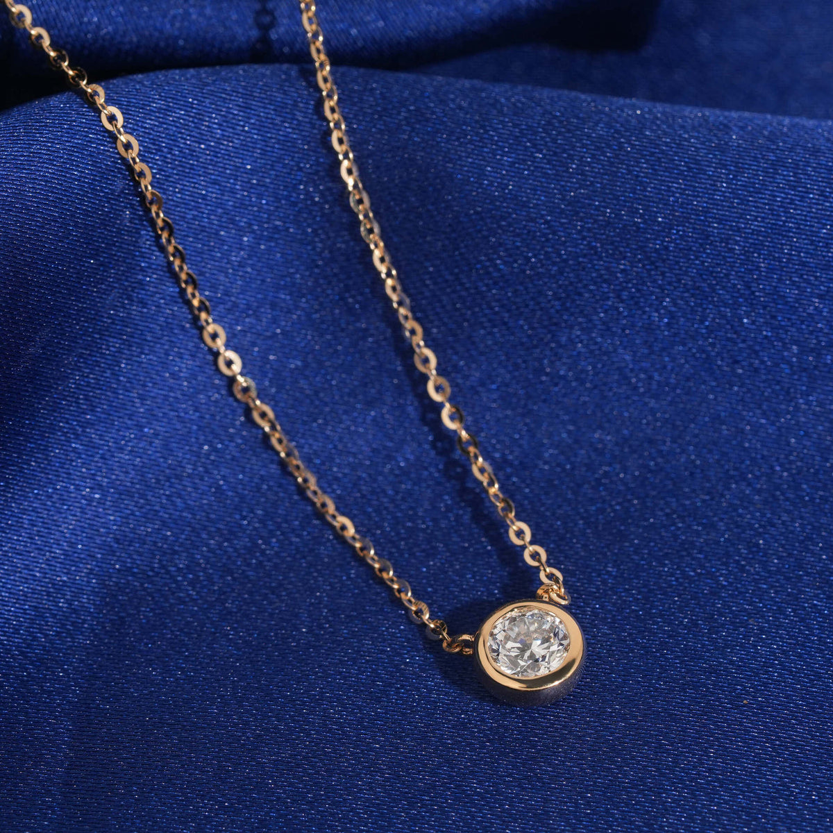 Angle view of 18k rose gold 5mm bezel-set solitaire lab-grown diamond pendant necklace on blue fabric   