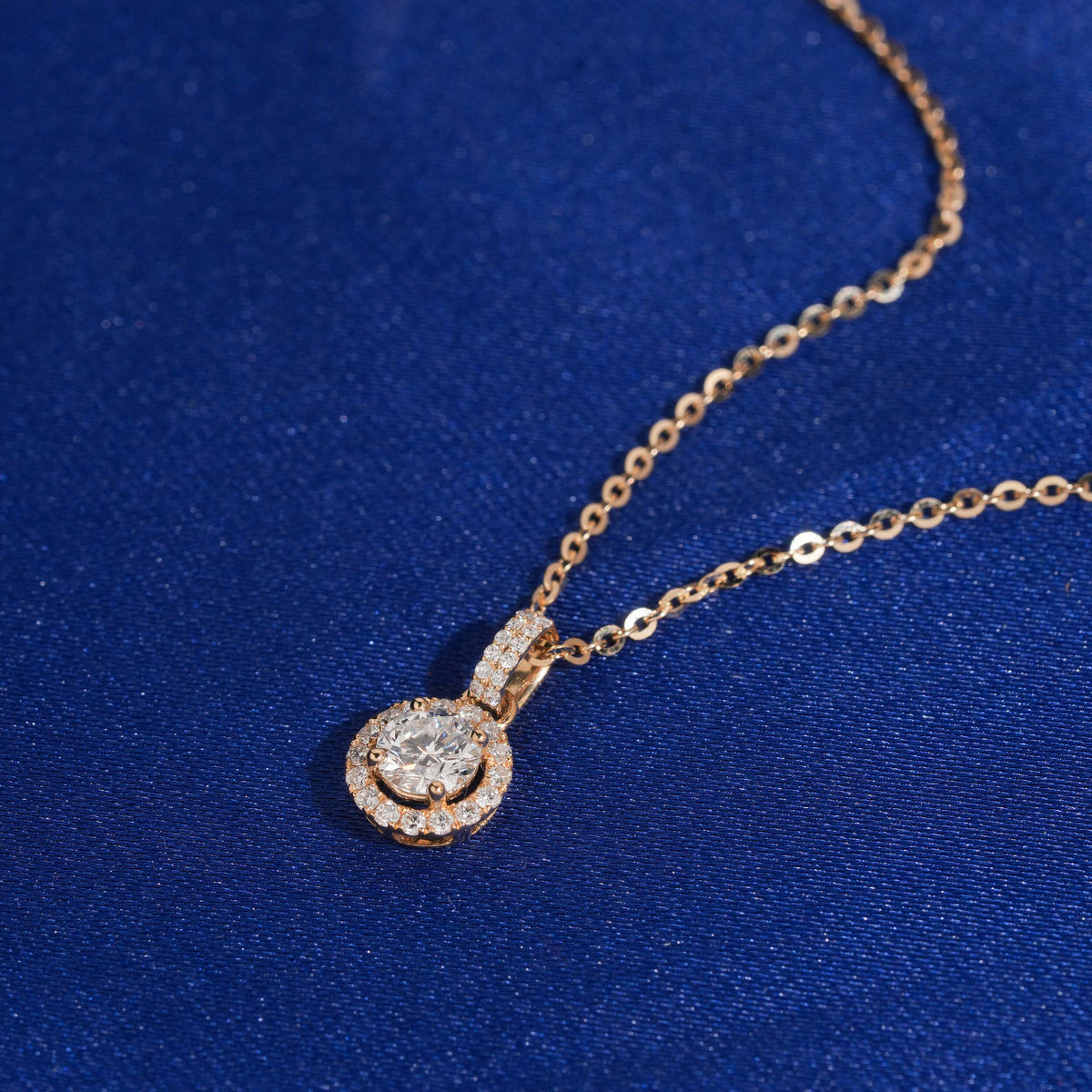 Angle view of 18k rose gold 5mm round halo lab-grown diamond clavicle pendant necklace on blue fabric
