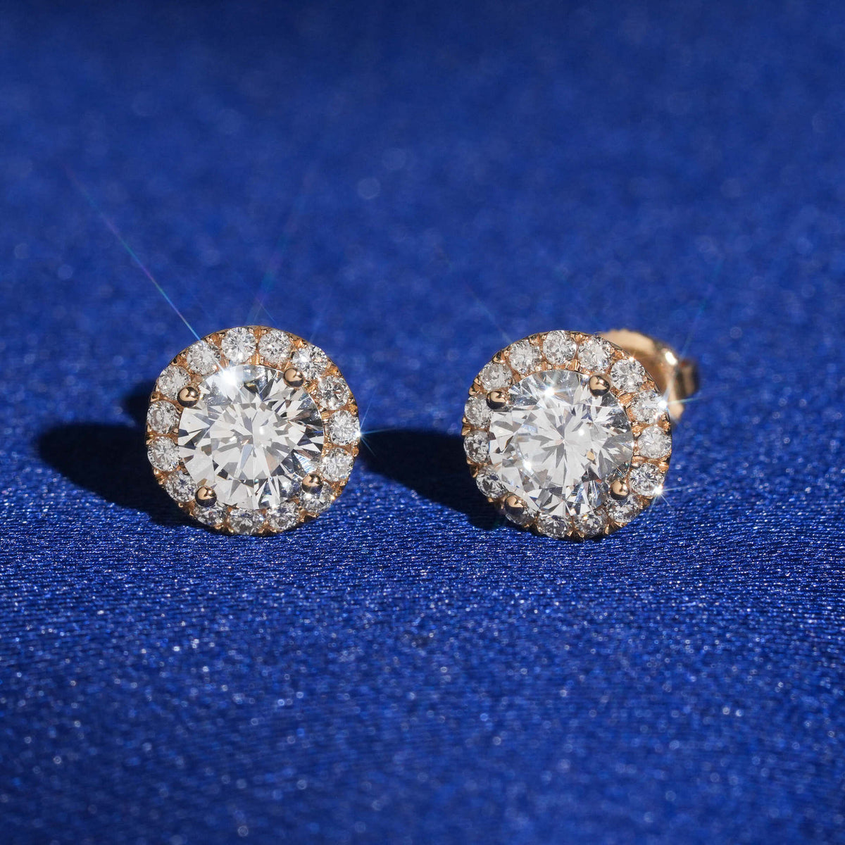 Pair of 18k rose gold 5mm round halo lab-grown diamond stud earrings standing on blue silk