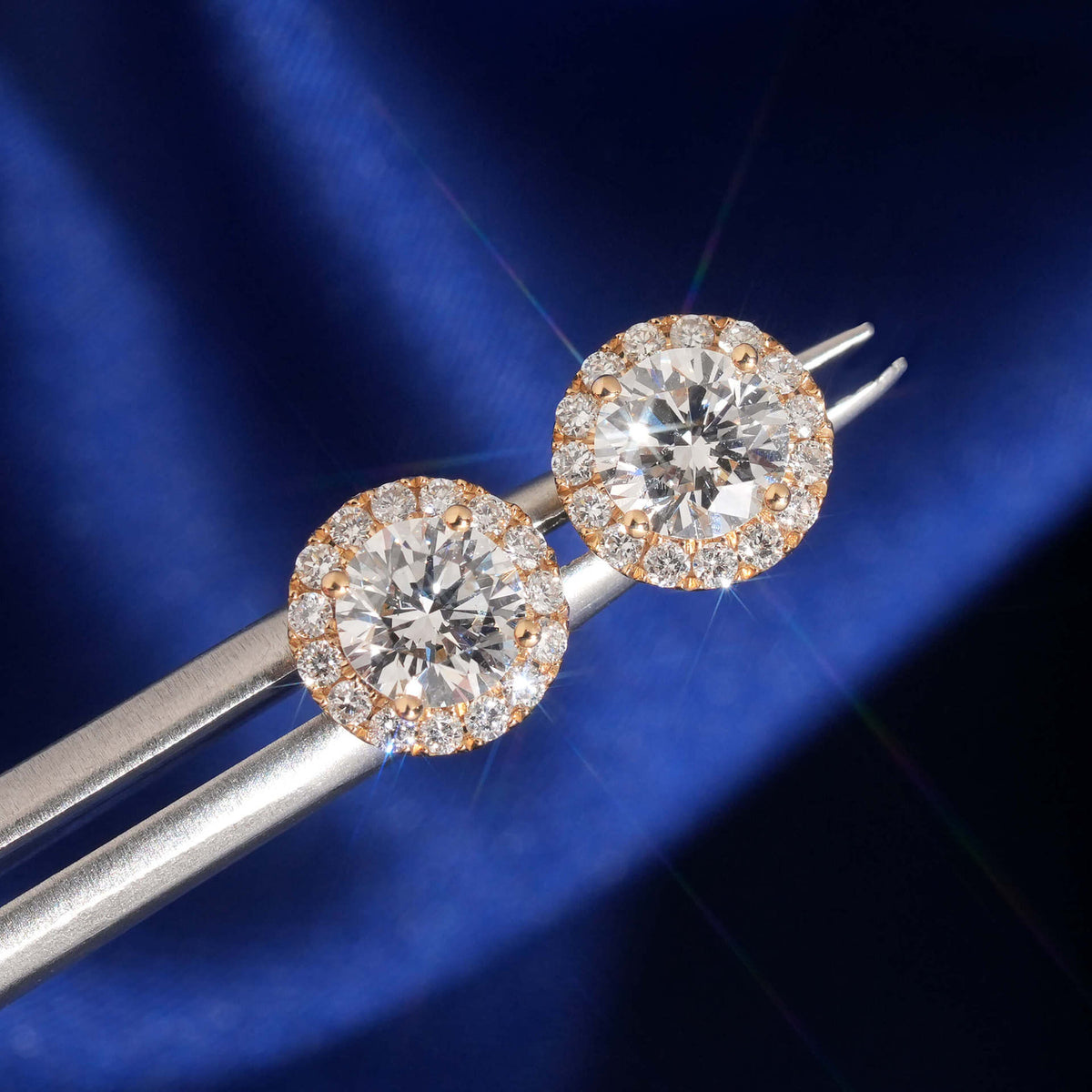 Tweezers holding 18k rose gold 5mm round halo lab-grown diamond stud earrings showing fire