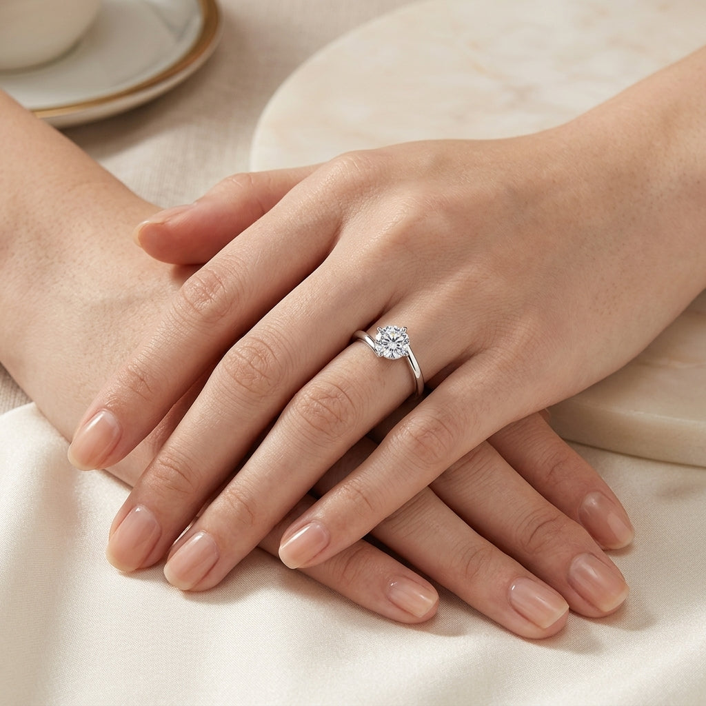 A silver ring with a round-cut clear gemstone worn on a woman's hand, resting on a soft cream fabric surface with a neutral background.