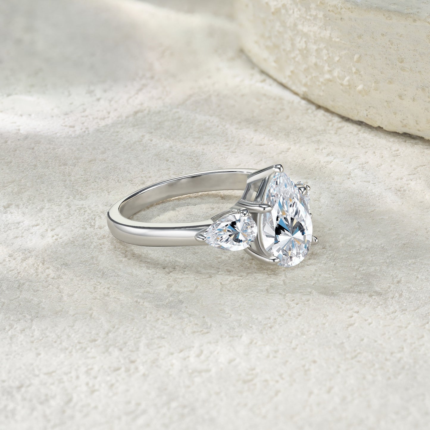Silver 3CT pear moissanite three-stone ring resting on a light textured background.