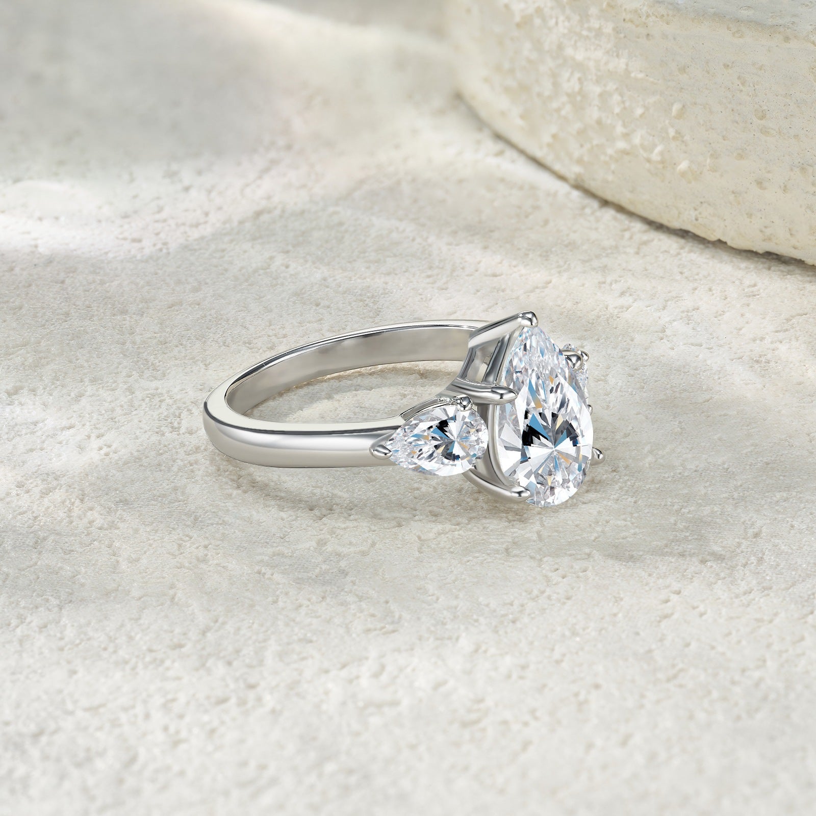 Silver 3CT pear moissanite three-stone ring resting on a light textured background.