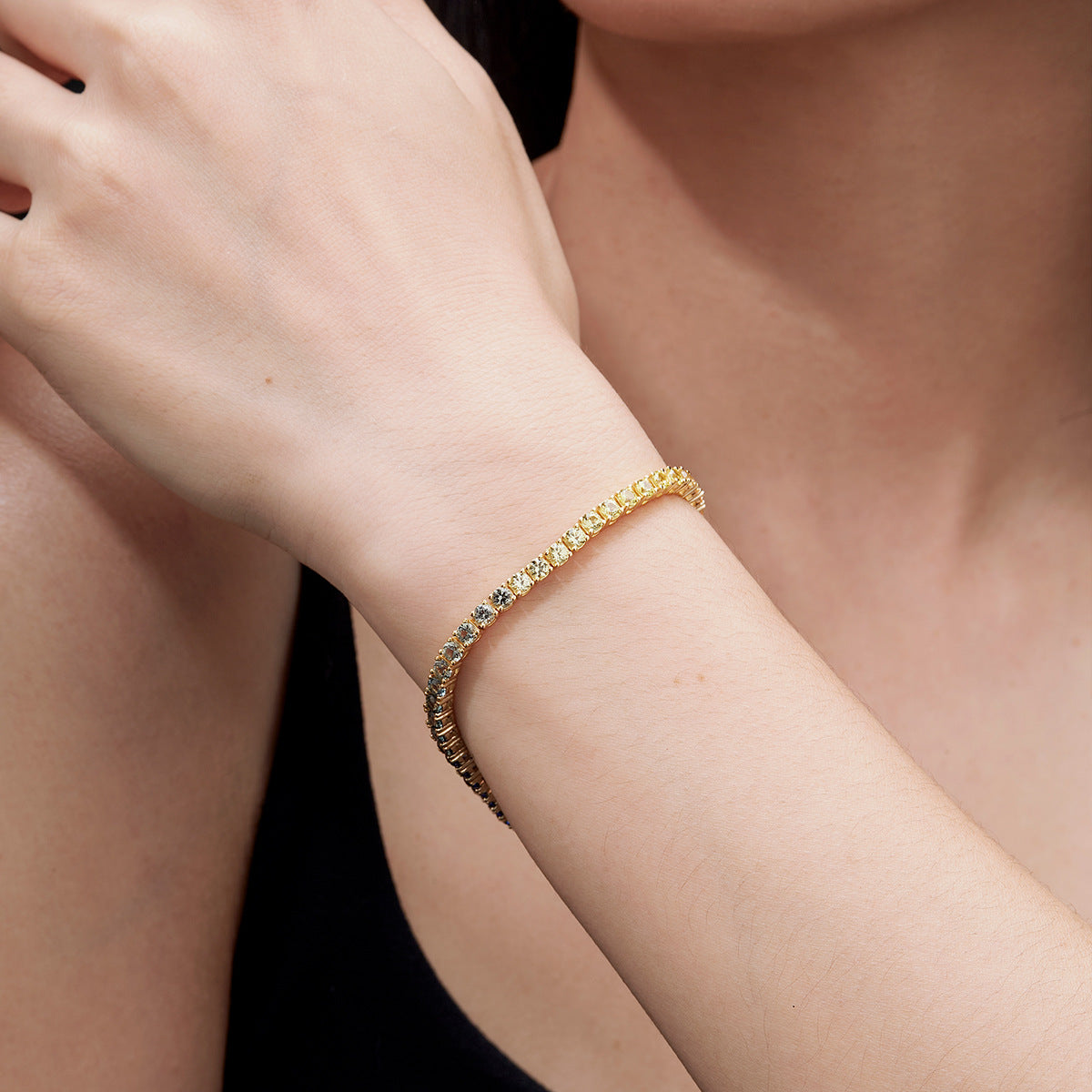 Model wrist closeup wearing gold 3mm round rainbow gradient moissanite tennis bracelet against dark backdrop