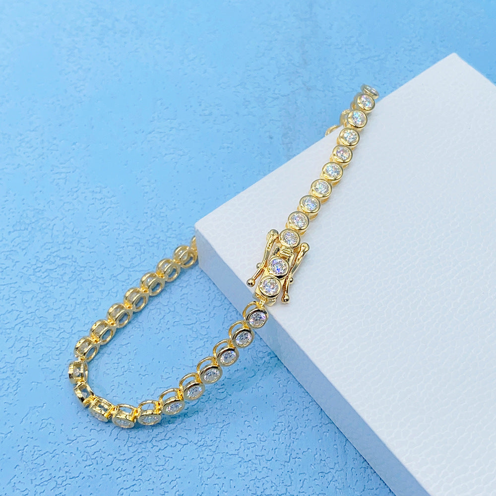 A gold tennis bracelet with clear round-cut stones, displayed on a white textured block against a light blue background.