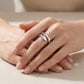 A close-up of a person's hands resting on a beige fabric surface, wearing a silver ring with a sculptural, layered geometric design.