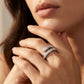 A close-up lifestyle shot of a model's hand wearing two silver rings, one featuring a row of clear pavé diamonds and the other a textured, diamond-like segmented design. The background is a soft, warm beige tone.