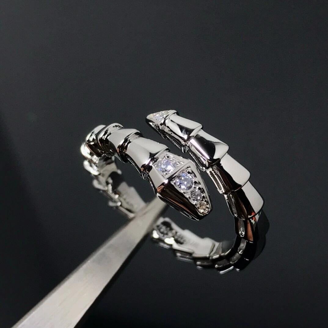 A silver-toned snake ring with clear gemstones, held in forceps against a dark reflective background.
