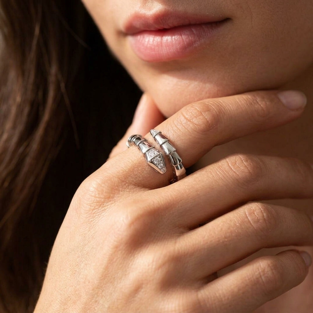 A close-up lifestyle shot of a model's hand resting near her chin, wearing two silver rings with textured, snake-like details and small clear stones.