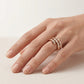 A close-up of a hand wearing a rose gold ring with multiple rows of small clear stones, set against a soft beige background.
