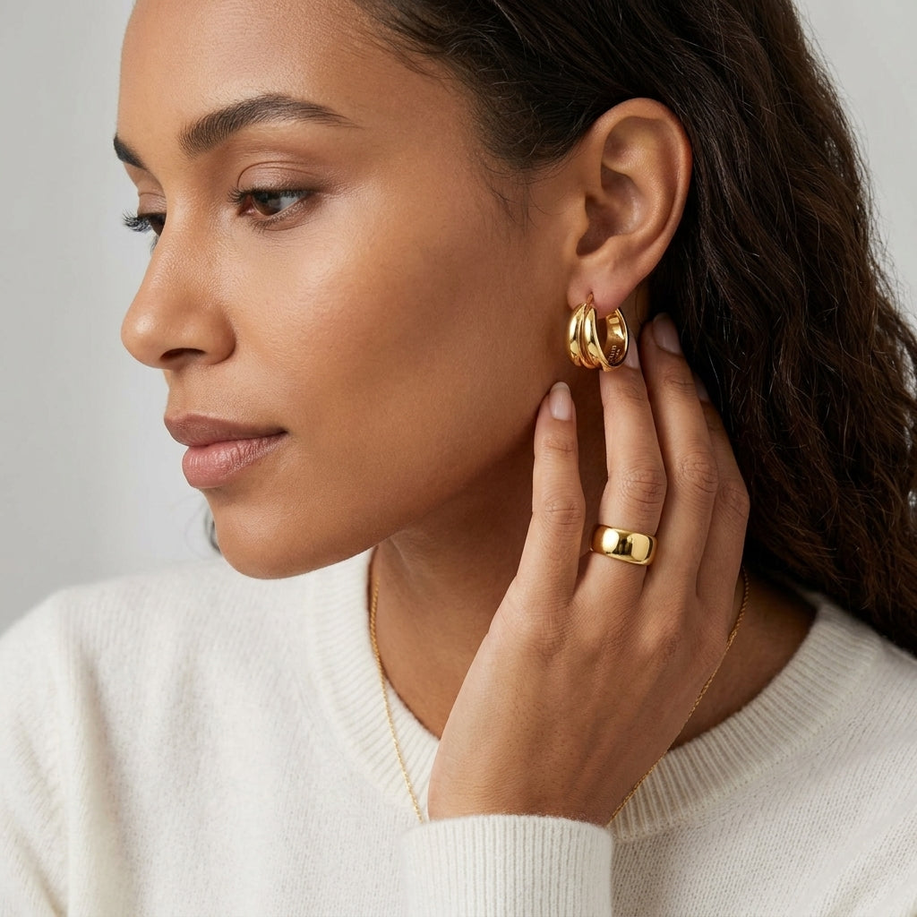 A close-up of a woman wearing gold hoop earrings and a gold ring, shown in profile against a soft light gray background.