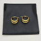 A pair of gold triple-band hoop earrings displayed on a black textured pouch against a light gray background.