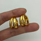 A pair of shiny gold triple-band hoop earrings held between fingers against a plain light background.