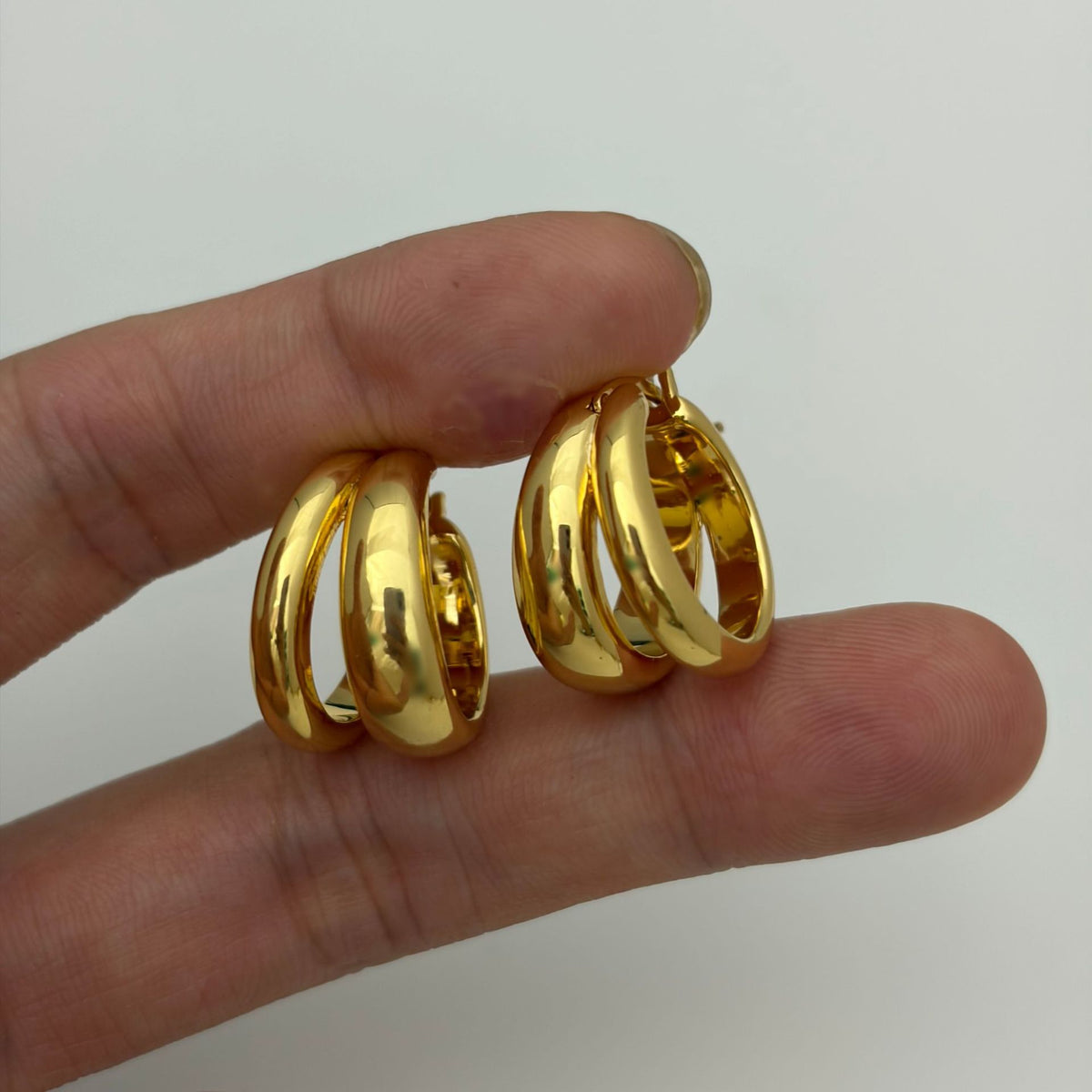 A pair of shiny gold triple-band hoop earrings held between fingers against a plain light background.