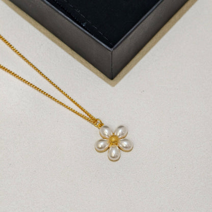 A gold chain necklace with a flower-shaped pendant made of white pearls and a gold center, displayed on a light beige surface next to a black jewelry box.