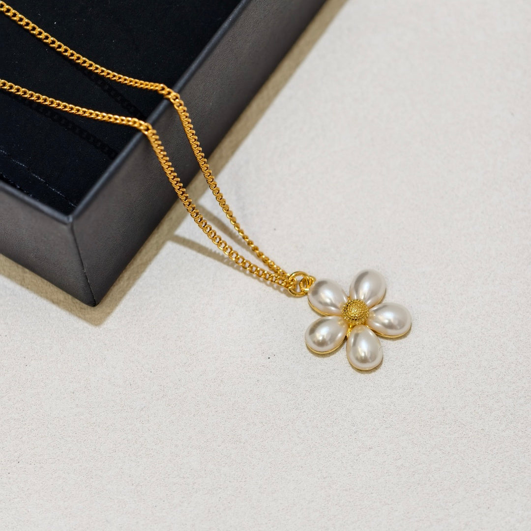 A gold chain necklace with a flower-shaped pearl pendant, displayed on a light beige surface next to a black jewelry box.