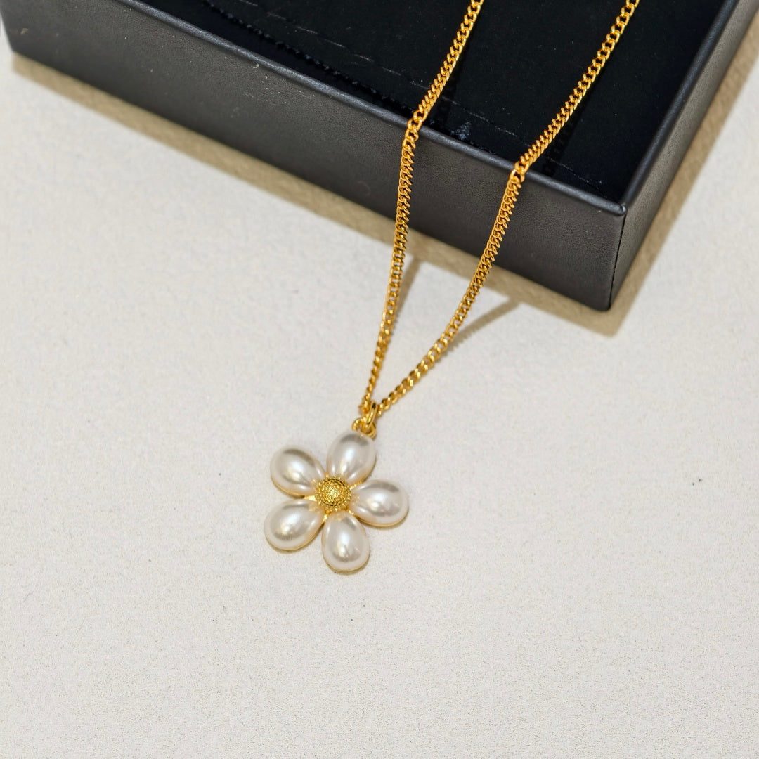 A gold chain necklace with a flower-shaped pendant made of five white pearls and a gold center, displayed on a light surface next to a black jewelry box.
