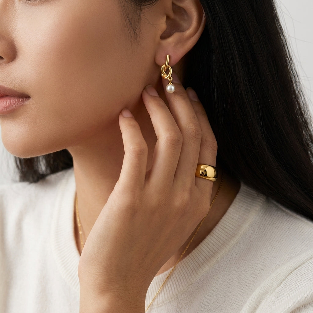 A close-up lifestyle shot of a woman wearing a delicate gold hoop earring with a small white pearl drop, paired with a gold ring and necklace.