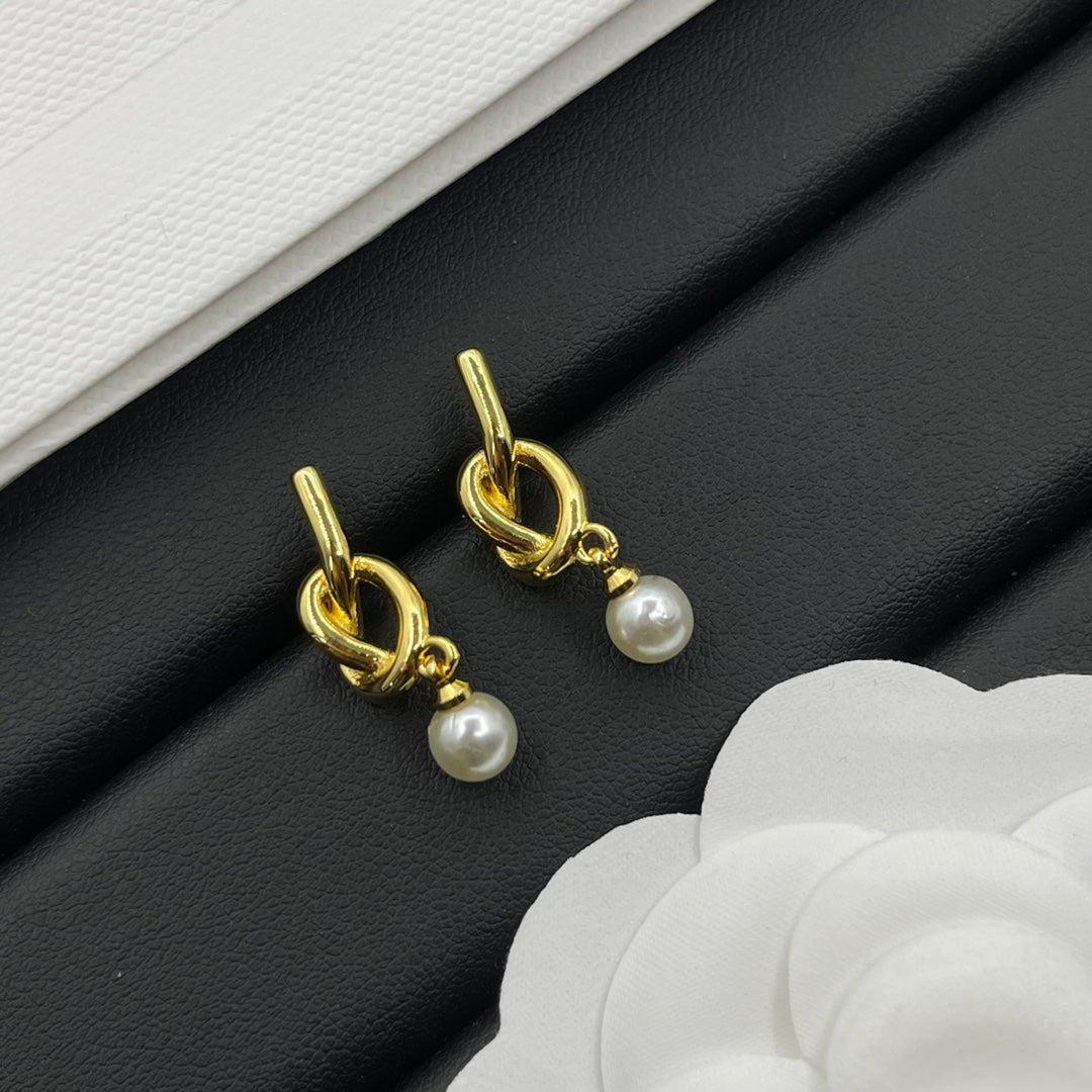 A pair of gold-toned drop earrings with a twisted loop at the top and a small white pearl dangling below, displayed on a black textured surface with white decorative paper accents.