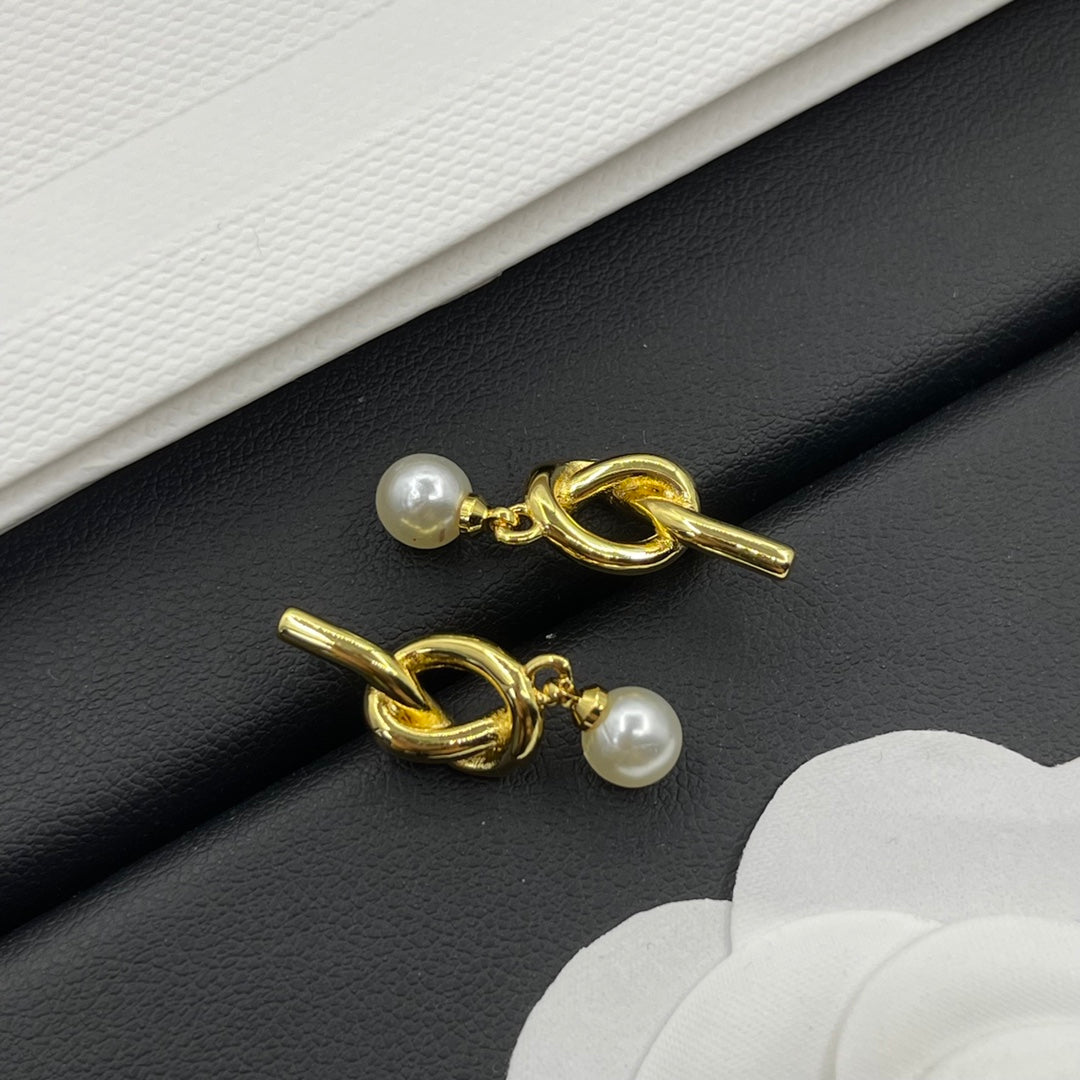 A pair of gold-toned drop earrings with a knotted loop design and a small white pearl dangling below, displayed on a black textured surface with white paper accents.