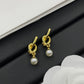 A pair of gold-toned drop earrings with a twisted knot design at the top and a small white pearl dangling below, displayed on a black textured surface with white decorative elements.