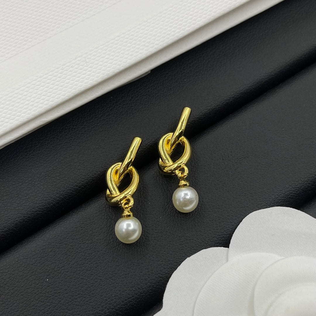A pair of gold-toned drop earrings with a twisted knot design at the top and a small white pearl dangling below, displayed on a black textured surface with white decorative elements.