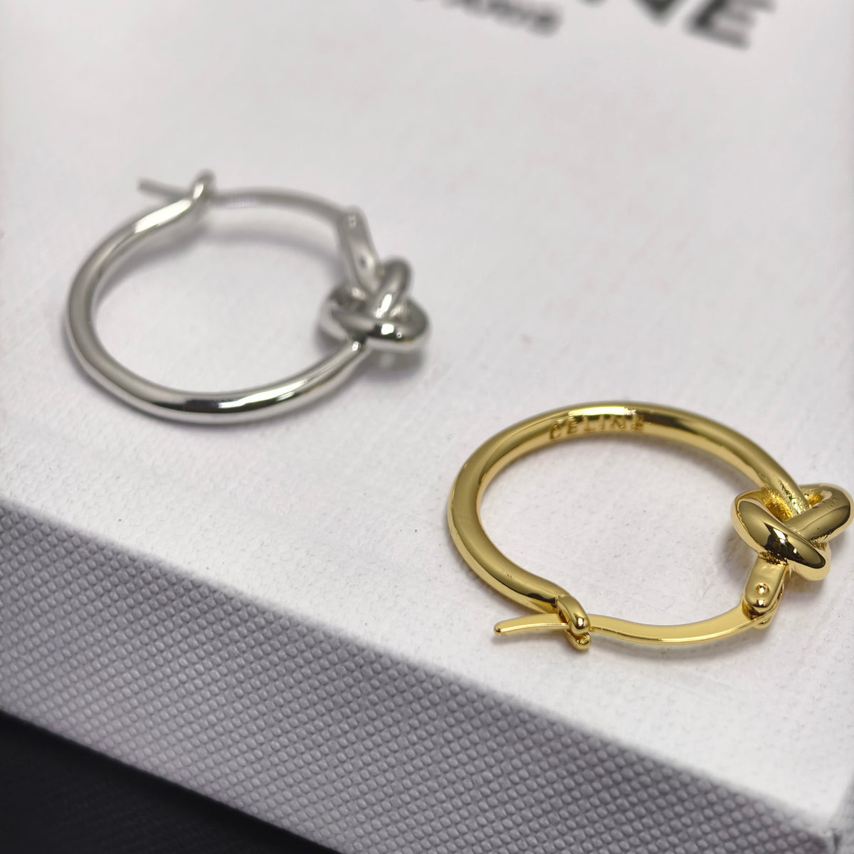 Two small hoop earrings with a knot detail, one silver-toned and one gold-toned, displayed on a textured white jewelry box.