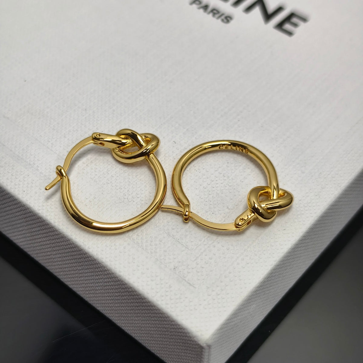 A pair of gold hoop earrings with a knotted detail at the front, resting on a white textured box with partial black text reading '.