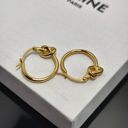 A pair of gold hoop earrings with a knotted detail at the front, resting on a white textured box with partial black text reading '.