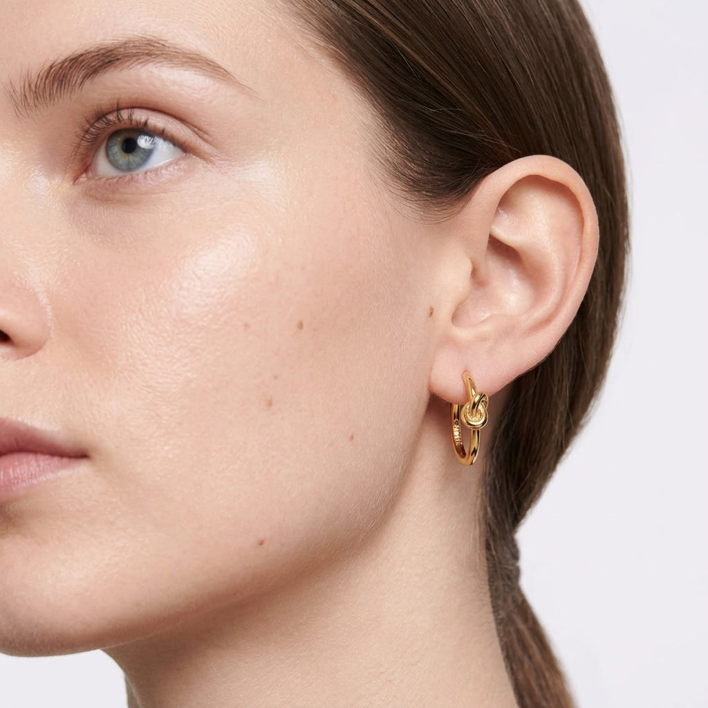 A close-up of a model's ear wearing a pair of small gold hoop earrings with a knotted detail, set against a clean white background.