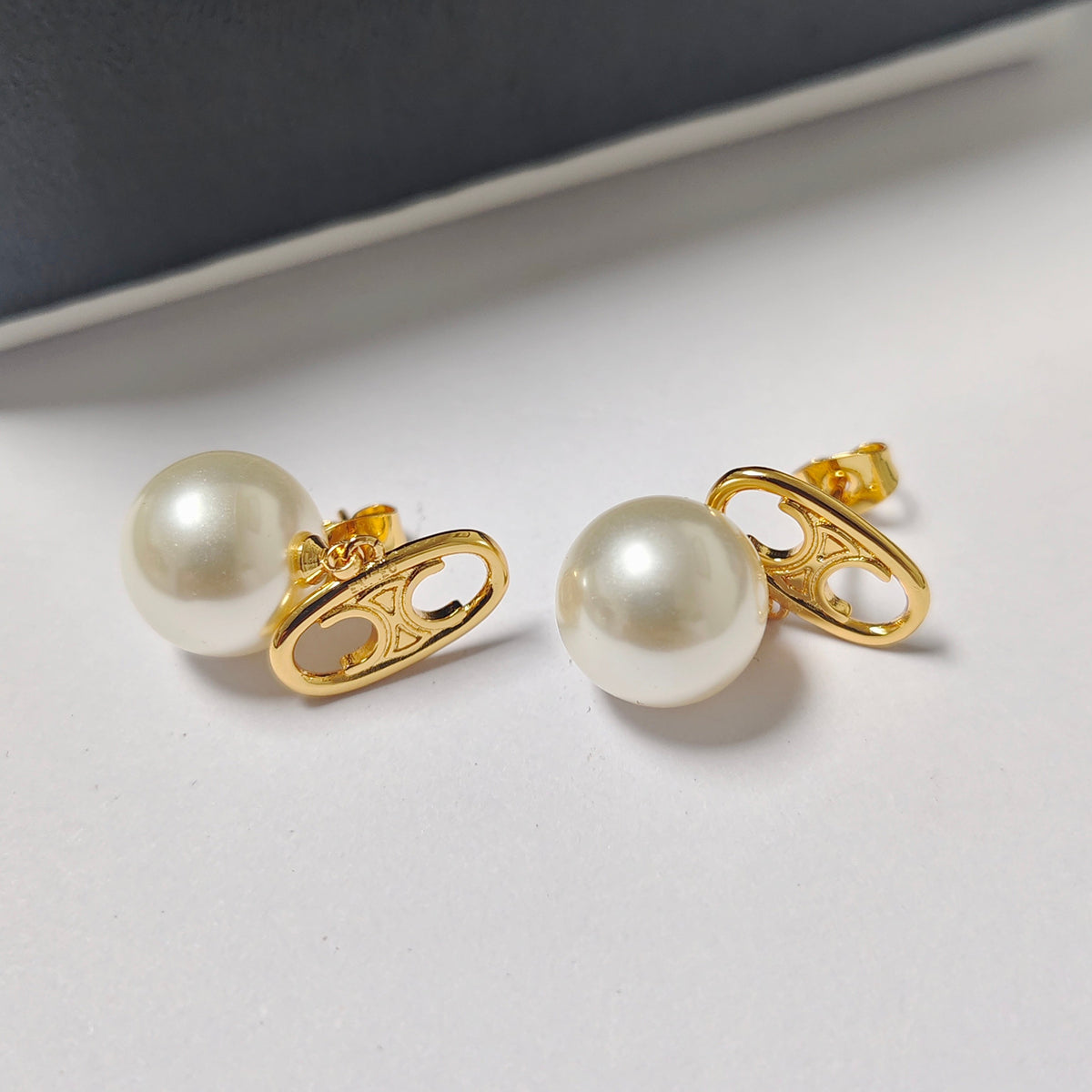 A pair of gold safety-pin stud earrings with white pearl centers, displayed on a clean white surface with a dark gray background.