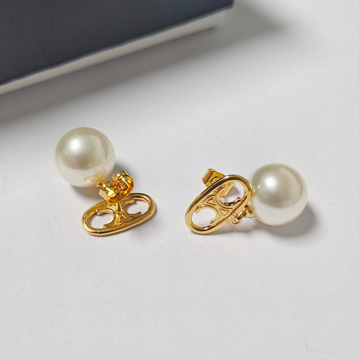 A pair of gold stud earrings with round white faux pearls and heart-shaped gold backings, displayed on a light gray surface.