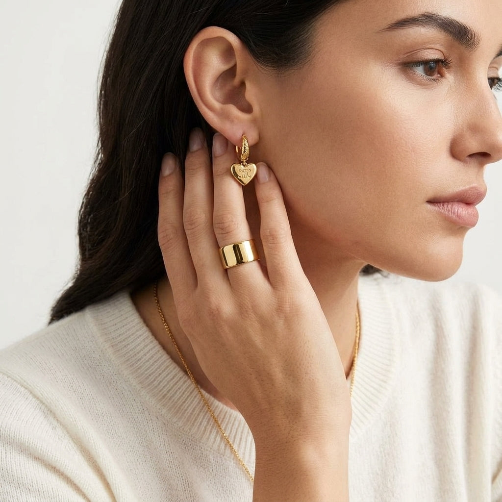 A close-up lifestyle shot of a woman wearing a small gold heart-shaped hoop earring and a wide gold ring, with a delicate gold chain necklace visible against a clean white background.
