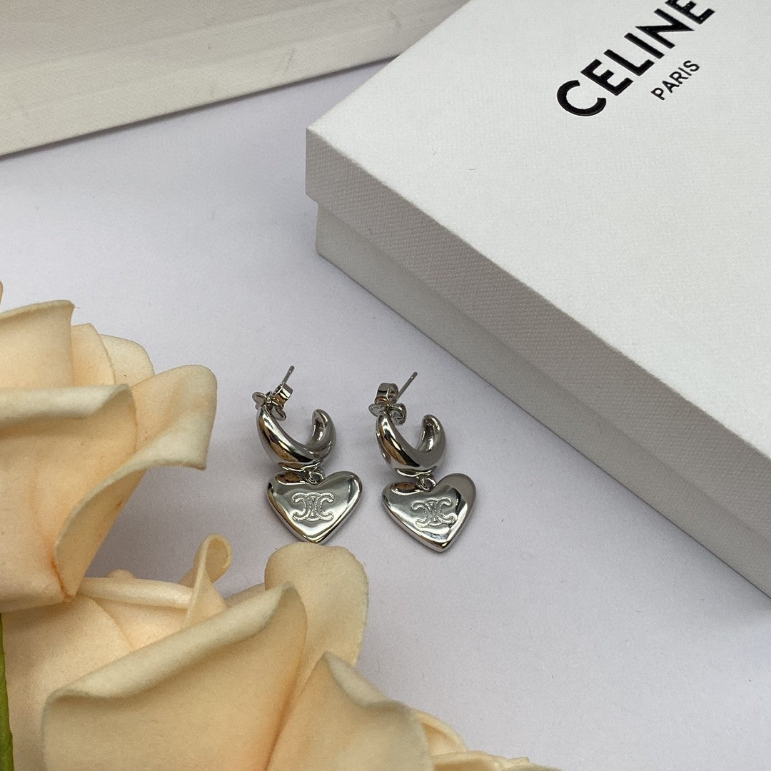 A pair of silver huggie hoop earrings with dangling heart charms engraved with the Celine logo, displayed next to a white Celine Paris box and cream-colored rose petals.