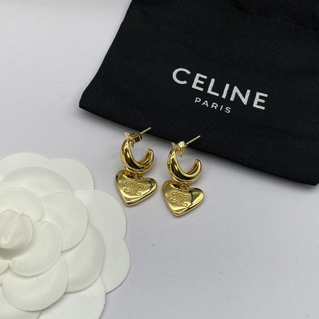 A pair of gold-tone Celine earrings with crescent and heart charms, displayed next to a black Celine Paris dust bag and a white flower prop on a light gray background.
